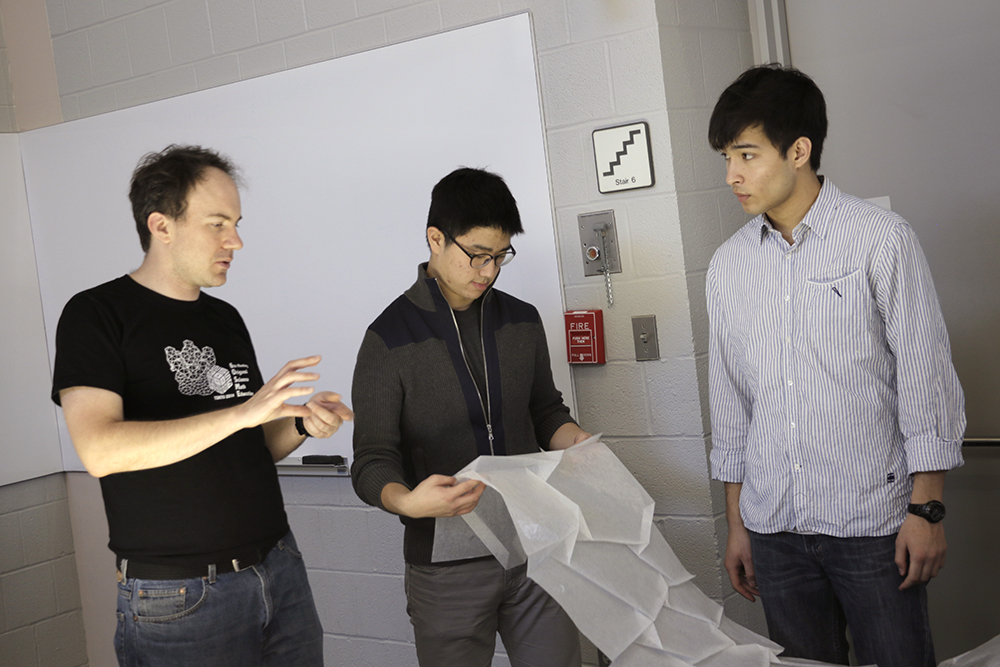 Three individuals on the Origami Cosmic Web team talk while examining folded tracing paper